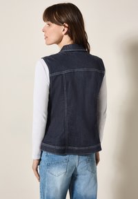 Denimväst i mörkblått med vit sömnad, krage och inga ärmar, ihop med ljusblå jeans. Bakvy, jämn yta, avslappnad design.
