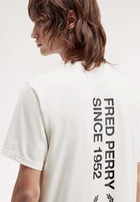 Weißes Baumwoll-T-Shirt mit einem vertikalen schwarzen Druck "FRED PERRY SINCE 1952" auf dem Rücken, rundem Halsausschnitt und kurzen Ärmeln.