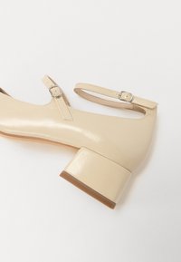 Beiga lackläder Mary Jane-skor med rund tå, ankelrem och blockklack, med en texturerad yta och spänndetaljer.