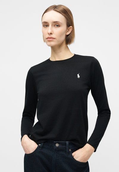 Γυναίκα με κοντά ξανθά μαλλιά, φορούσα μαύρο μπλουζάκι με μακριά μανίκια και λευκό λογότυπο, και σκούρο μπλε τζιν, στέκεται με τα χέρια στις τσέπες.