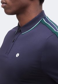 Κοντινό πλάνο ενός ατόμου που φοράει μπλε κοντομάνικο πόλο με πράσινη και λευκή ριγέ λεπτομέρεια στον ώμο και ένα μικρό λευκό λογότυπο στο στήθος.