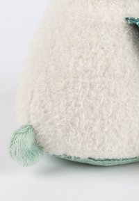 Cream-colored plush toy with a soft, fluffy texture, featuring a rounded green tail and a flat base with green fabric accents.