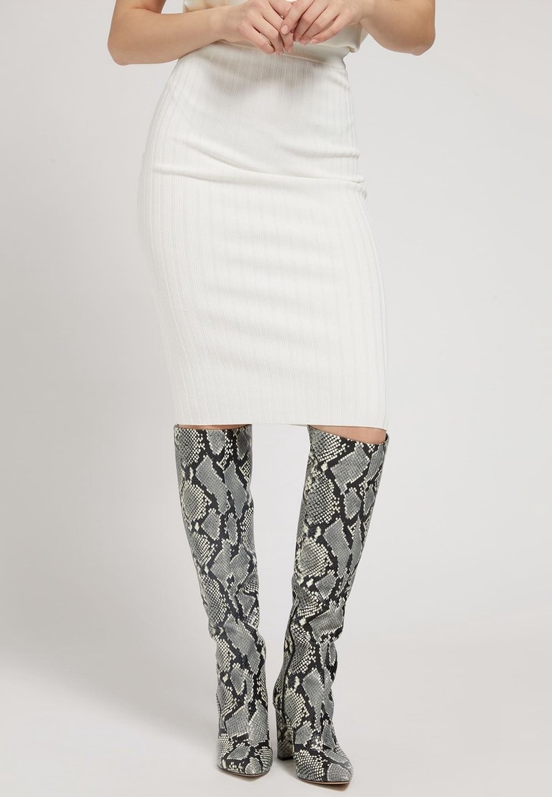Femme portant une jupe midi blanche côtelée associée à des bottes grises à motifs peau de serpent montant jusqu'aux genoux, debout devant un fond uni.