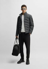 Puffjacka i mörkgrått med hög krage, matchad med en vit tröja, svarta tapered byxor och svarta sneakers. Håller i en svart duffelväska.