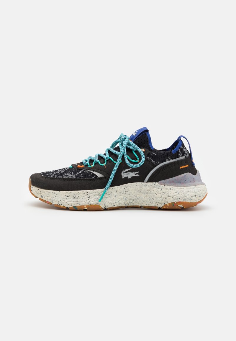 Zapatilla deportiva negra con parte superior texturizada, cordones turquesa y suela moteada en blanco y naranja. Presenta un logotipo prominente en el costado.