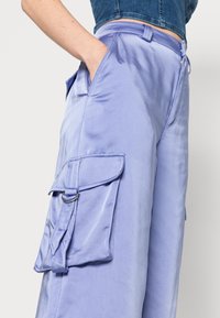 Calças cargo de satin lilás claro com um bolso lateral, costura visível e passante para cinto. Textura suave e corte largo.
