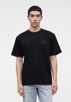 Giovane uomo con capelli neri e corti che indossa una t-shirt nera Lacoste e jeans azzurri, in piedi davanti a uno sfondo bianco semplice.