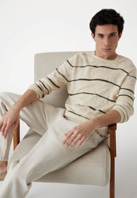 Pull beige à rayures avec des lignes vert foncé, porté avec un pantalon de couleur claire. Le mannequin est assis sur une chaise, montrant une posture décontractée et les mains.