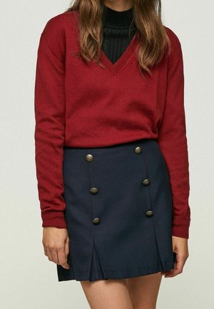 Femme portant un pull rouge à col en V par-dessus un haut noir, associé à une jupe bleu marine ornée de deux rangées verticales de boutons décoratifs.