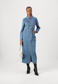 Denimmekko, jossa on napitettu etuosa, pitkät hihat, kaulus ja etutaskut. Vaaleansininen väri ja suora leikkaus. Yhdistetty polvipituisien mustien saappaiden kanssa.