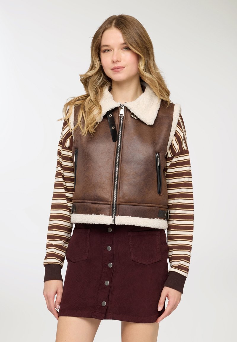 Gilet in pelle marrone con collo in shearling crema, cerniera frontale e tasche laterali. Abbinato a una camicia a righe marrone e bianca e una gonna bordeaux.