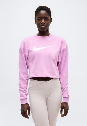 Žena v ružovej krátkej mikine s bielym logom Nike a béžových legínach s vysokým pásom na jednofarebnom pozadí.