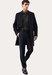 Marineblauer Wollmantel über einem dunkel gemusterten Hemd, kombiniert mit maßgeschneiderten dunklen Hosen und schwarzen Lederschuhen. Klare Linien und eine taillierte Silhouette.