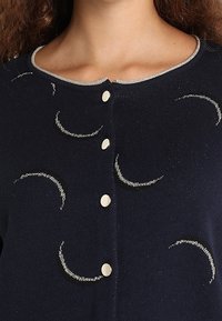 Granatowy kardigan z wzorem srebrnych półksiężyców, okrągłym dekoltem z metalicznym obszyciem oraz pięcioma białymi guzikami z przodu.