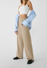 Pantalons larges beiges associés à un top court blanc et une chemise oversize rayée bleue, avec fermeture à boutons et poches latérales.
