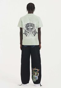 Una camiseta de manga corta verde claro presenta un diseño de un tigre negro en la parte de atrás, combinada con pantalones cargo negros que muestran acentos gráficos coloridos.