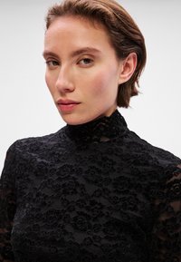 Μαύρη δαντελένια μπλούζα με μακριά μανίκια, που διαθέτει ψηλή λαιμόκοψη, λουλουδάτα μοτίβα και υφή, με διάφανο ύφασμα στα μανίκια.