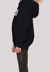 Sudadera negra con un bolsillo frontal, que presenta un diseño gráfico colorido. El modelo la combina con leggings verde oliva que tienen un detalle de malla.