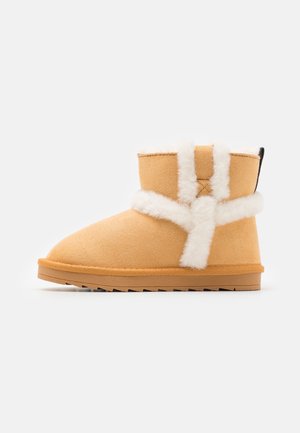 Hellbrauner Wildleder-Stiefelette mit weißem flauschigem Besatz, flacher Gummisohle und Zuglasche an der Rückseite, auf weißem Hintergrund dargestellt.