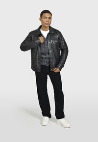 Čierna kožená bunda s predným zapínaním na gombíky, pod ňou štruktúrovaná látka, tmavé nohavice a biele tenisky. Model stojí proti neutrálnemu pozadiu.