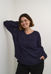 Tamno plava, lagana, dugih rukava majica s V-izrezom, opuštenog kroja i teksturirane tkanine. Model pozira uz zid, ruku na bokovima.
