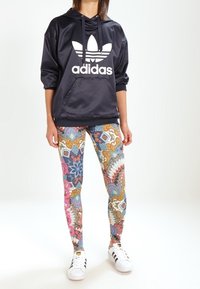 Marinblå satin hoodie med vitt Adidas-logga, kombinerad med färgglada mönstrade leggings med blommor och geometriska mönster, samt vita sneakers.