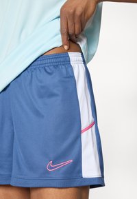 Αθλητικό σορτς Nike με μπλε ύφασμα, λευκές πλευρές, ροζ λεπτομέρειες, ελαστική μέση και πλαϊνή τσέπη.