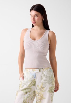 Kobieta o długich ciemnych włosach, ubrana w bezrękawnik beżowy dzianinowy top i jasne spodnie z zielono-niebieskim wzorem liści, stoi z rękami w kieszeniach.