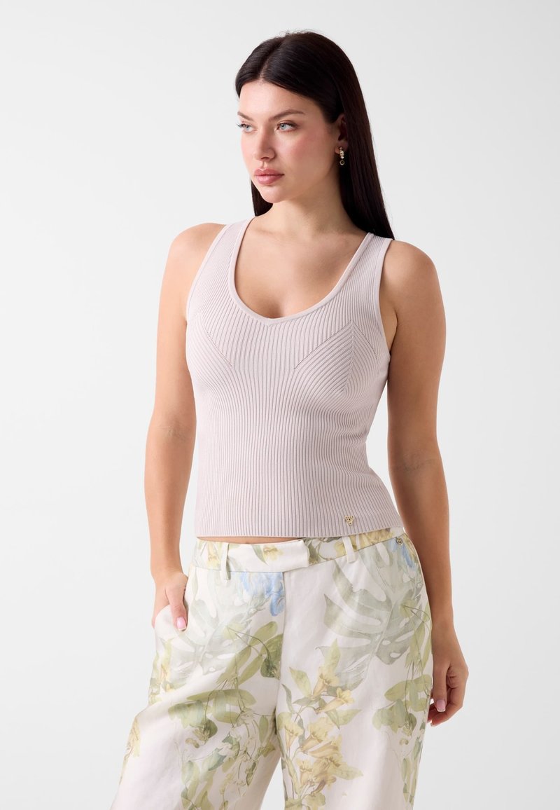 Femme aux longs cheveux foncés portant un haut beige côtelé sans manches et un pantalon clair avec un motif de feuilles vertes et bleues, debout les mains dans les poches.