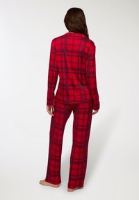 Donna in piedi a piedi nudi, con una camicia a maniche lunghe a quadri rossi e blu scuri e pantaloni abbinati, rivolta verso l'ingiù su uno sfondo bianco.