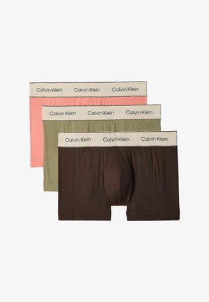 Muskelbepackter Mann steht barfuß mit verschränkten Armen da und trägt olivgrüne Calvin Klein Boxershorts vor einem schlichten weißen Hintergrund.