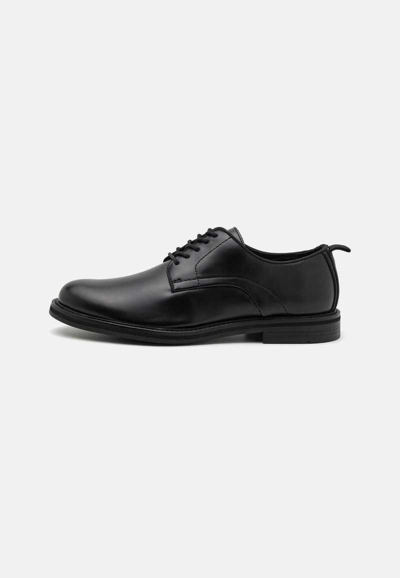 Chaussure habillée pour hommes en cuir noir poli avec des lacets et un petit talon, vue de profil sur fond blanc.