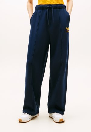 Osoba odjevena u široke sportske hlače tamnoplave boje s vezicom u struku, ruke u džepovima, bijele tenisice i gornji dio u boji gorušice.