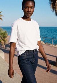 Jeune femme marchant le long d'une promenade en bord de mer, portant un t-shirt blanc et un jean bleu foncé sous un ciel bleu clair.