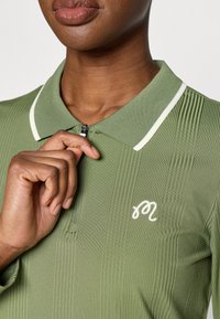 Zaļa ridēta polo krekls ar baltu apkakli un rāvējslēdzēju uz kakla. Blakus apkaklei redzams iešūts balts logo.