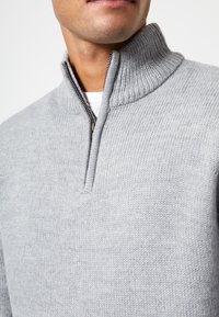 Homme portant un pull en tricot gris clair avec un col montant côtelé et une fermeture éclair partiellement ouverte sur une chemise blanche, gros plan.