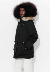 Parka noire avec une capuche doublée en fourrure, fermeture à boutons sur le devant, poches latérales et cordon de serrage ajustable à la taille pour un ajustement sur mesure.