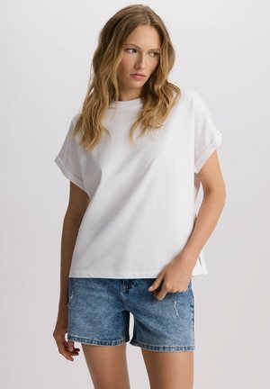 Biała koszulka z krótkim rękawem o luźnym kroju, z okrągłym dekoltem i podwiniętymi mankietami, w połączeniu z niebieskimi szortami dżinsowymi z delikatnymi przetarciami.