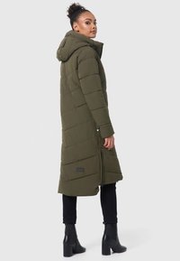 Manteau matelassé vert olive avec capuche, présentant un motif matelassé diagonal, des zips latéraux et un patch logo ; porté avec un pantalon noir et des bottes à talons.