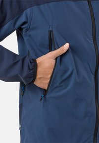 Hand gestoken in een met rits afsluitbare zak van een blauw- en marineblauwe jas met elastische manchetten en zwarte ritsdetails.