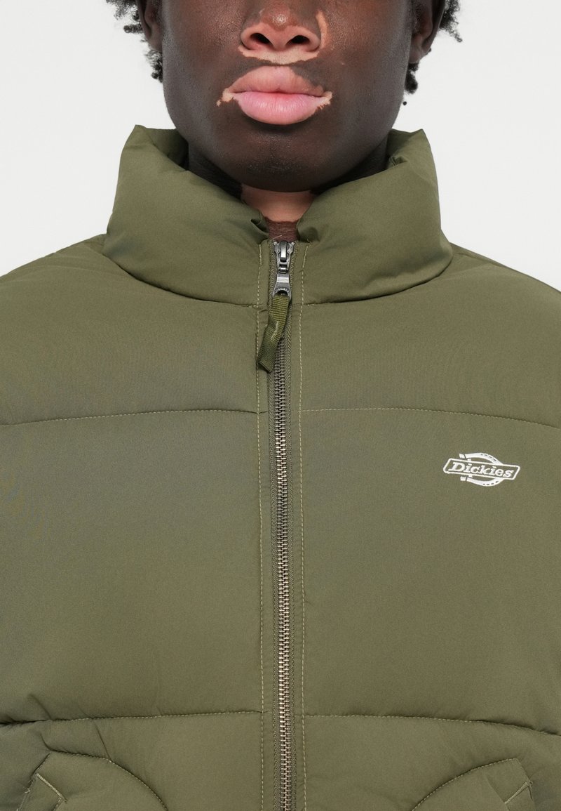 Olivgrüne gepolsterte Jacke mit hohem Kragen, Frontreißverschluss und gesticktem Logo auf der linken Brust. Strukturierter Stoff mit gestepptem Muster.