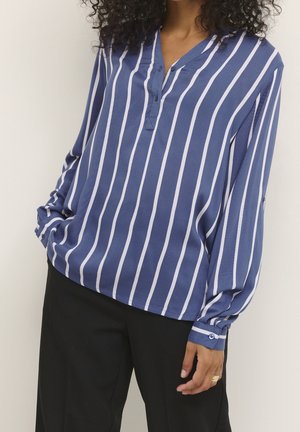 Kobieta ubrana w niebiesko-białą koszulę z pionowymi paskami na długim rękawie oraz czarne spodnie, stojąca z jedną ręką w kieszeni, a drugą luźno opuszczoną.