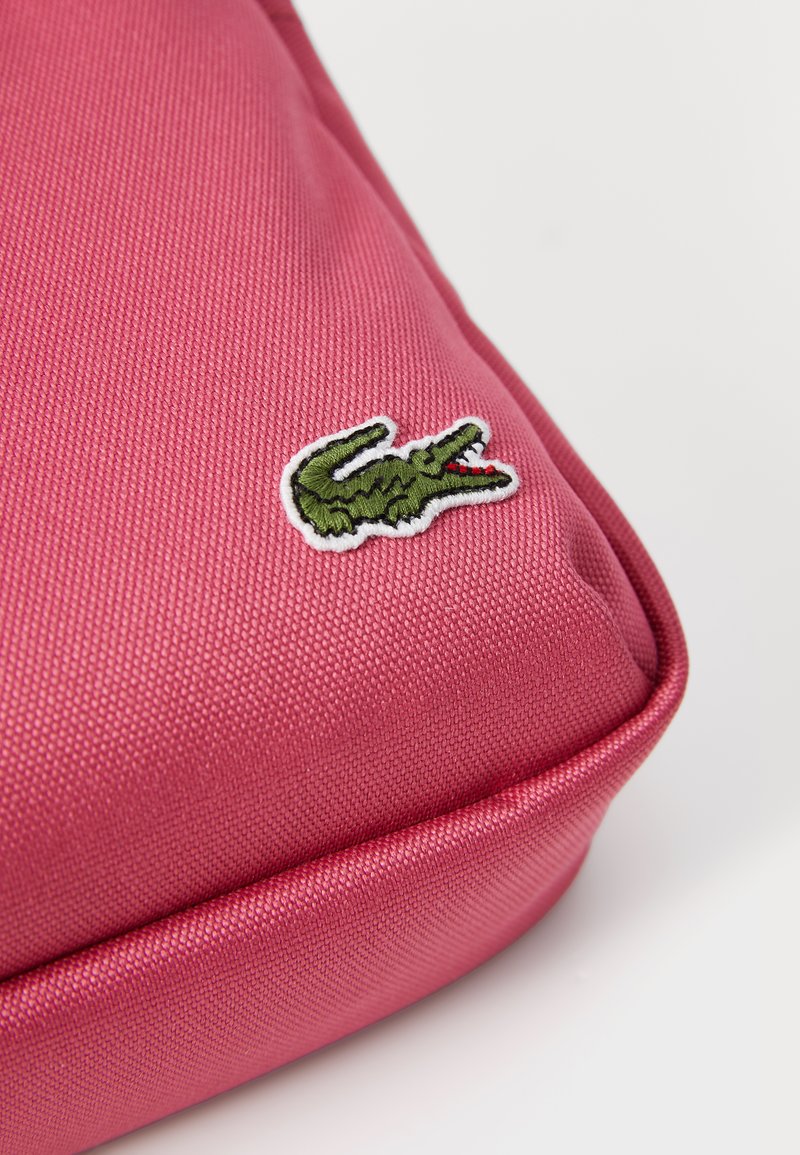 Poche en tissu rose avec une surface texturée, présentant un logo de crocodile brodé en vert. Bords arrondis et couture contrastée.