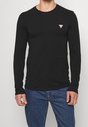 Homme portant une chemise noire ajustée à manches longues avec un petit logo triangulaire Guess sur la poitrine et un jean bleu sur un fond uni.