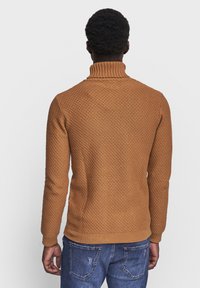 Pull à col roulé de couleur ambre chaude, avec un motif tricoté texturé et des poignets et un ourlet côtelés, associé à un jean bleu.