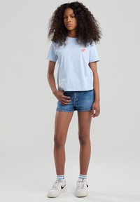 T-shirt azzurro in cotone a maniche corte con logo rosso, abbinato a pantaloni corti di denim e sneakers bianche con logo swoosh nero.