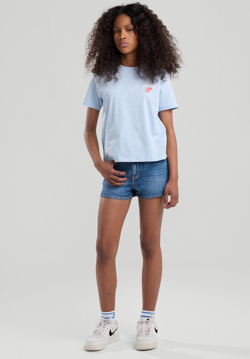 T-shirt azzurro in cotone a maniche corte con logo rosso, abbinato a pantaloni corti di denim e sneakers bianche con logo swoosh nero.