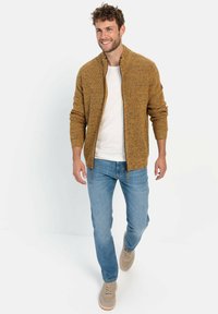 Gestrickter Reißverschluss-Pullover in einer strukturierten Senffarbe, kombiniert mit einem weißen T-Shirt und blauen Jeans. Hat einen Kragen und gerippte Bündchen.