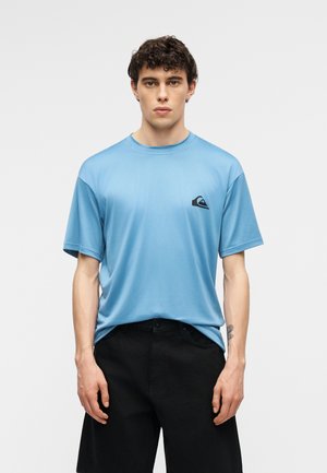 Junger Mann mit lockigem Haar, der ein hellblaues Kurzarmhemd mit schwarzem Logo auf der Brust und schwarze Hose trägt, vor einfarbigem Hintergrund stehend.