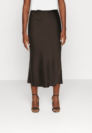 Žena v tmavě hnědé midi sukni, bílém topu a černých sandálech na vysokém podpatku s páskem kolem kotníku, stojící před jednobarevným pozadím.